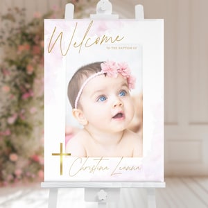 May include: Welcome to the Baptism of Christina Leanna sign with a watercolor background and a photo of a baby girl wearing a pink headband and a pink flower crown. The sign has a gold cross and the baby's name in gold script.