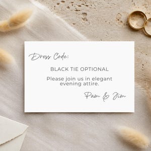 Może przedstawiać: Prostokątna biała karta z czarnym tekstem o treści Dress Code: BLACK TIE OPTIONAL. Please join us in elegant evening attire. Pam & Jim. Karta jest otoczona suszonymi beżowymi trawami oraz dwoma złotymi pierścionkami.