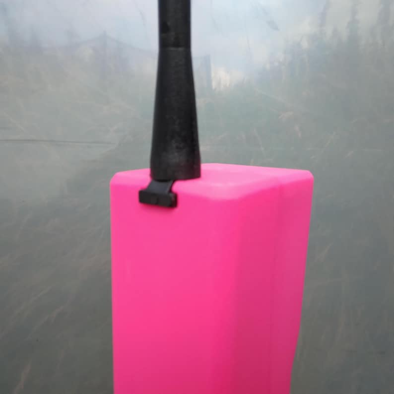 May include: A bright pink, rectangular object with a black handle and connector. The object appears to be a part of a larger tool or device. The background is a blurred, neutral tone.