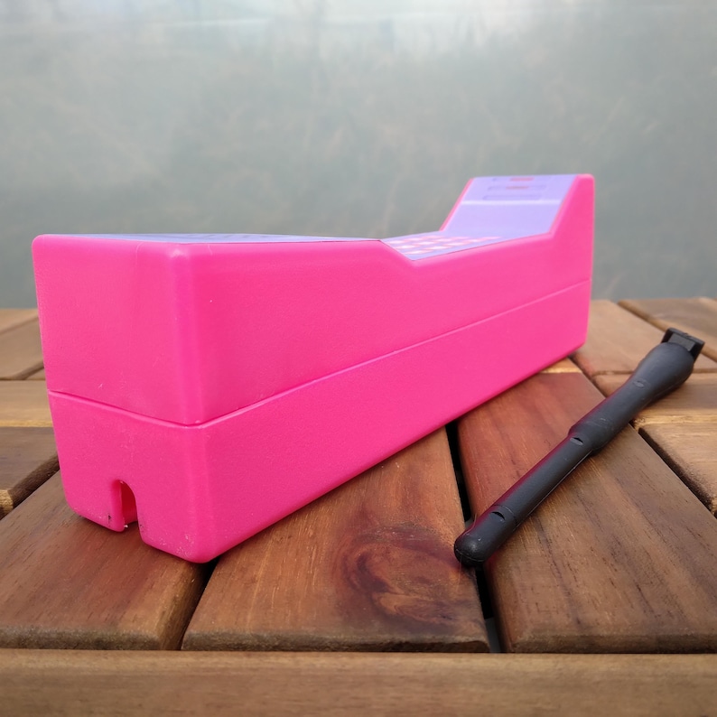 May include: A bright pink toy cash register with a purple top section and a black stylus. The register is rectangular and sits on a wooden surface. The stylus is cylindrical with a rounded tip.