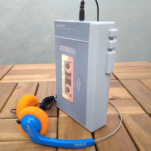 May include: A vintage gray Sony Walkman cassette player with a cassette tape design on the front, connected to orange and blue headphones. The player is plugged in and sits on a wooden surface.