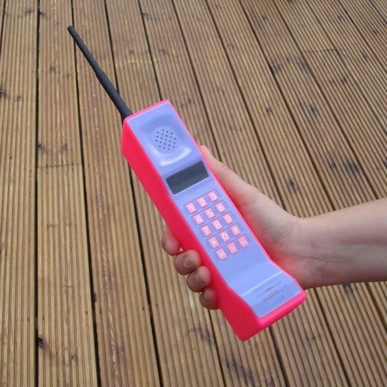 May include: A retro-style, oversized cell phone in a vibrant pink and lavender color scheme. The phone features a keypad with pink buttons, a speaker, and a black antenna. The phone is held in a hand.