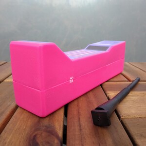 May include: A bright pink toy cash register with a checkerboard design on the top and a black stylus. The register is rectangular with a sloped top and is sitting on a wooden surface.