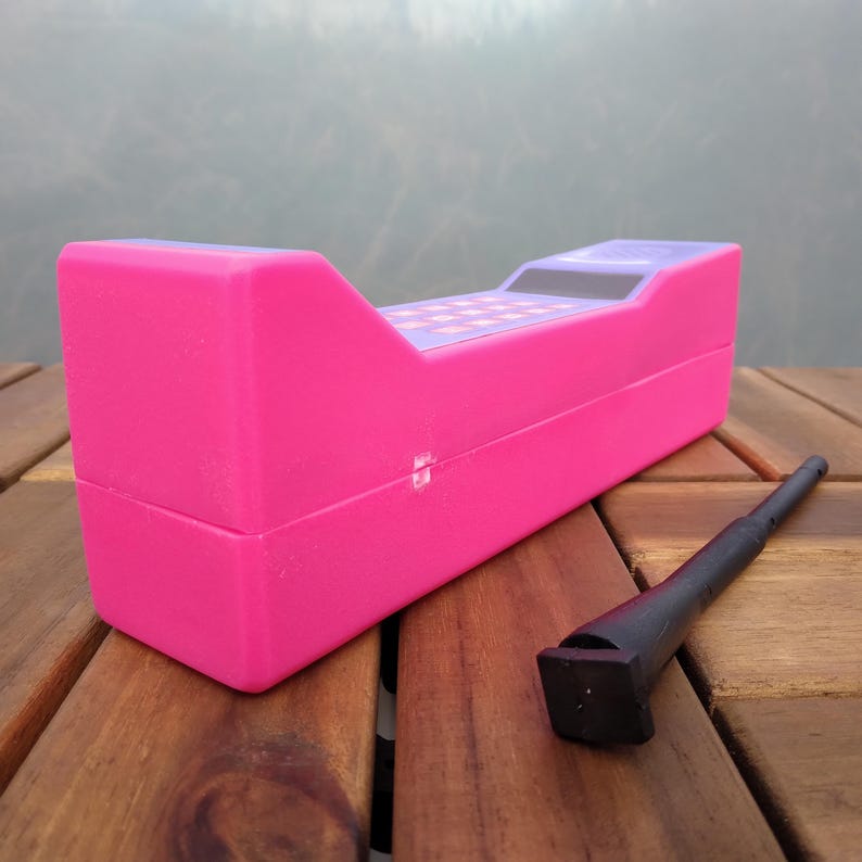 May include: A bright pink toy cash register with a checkerboard design on the top and a black stylus. The register is rectangular with a sloped top and is sitting on a wooden surface.