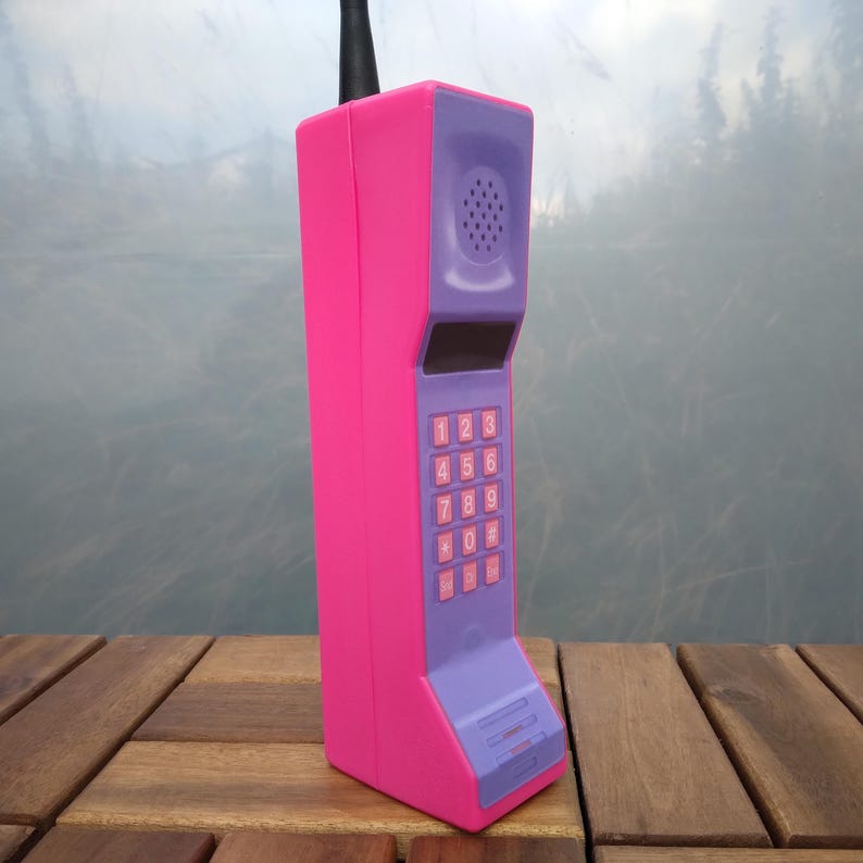 May include: A retro-style, oversized cell phone in bright pink and purple. The phone features a keypad with numbers, a speaker, and a black antenna. The design is reminiscent of early mobile phones from the 1980s.