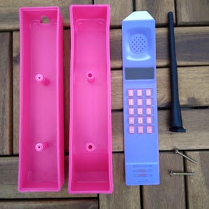 May include: A disassembled toy cellular phone with a purple keypad and antenna, pink casing, and two screws. The phone has a white top, a speaker, and a number pad with pink buttons. The pink casing is in two parts.