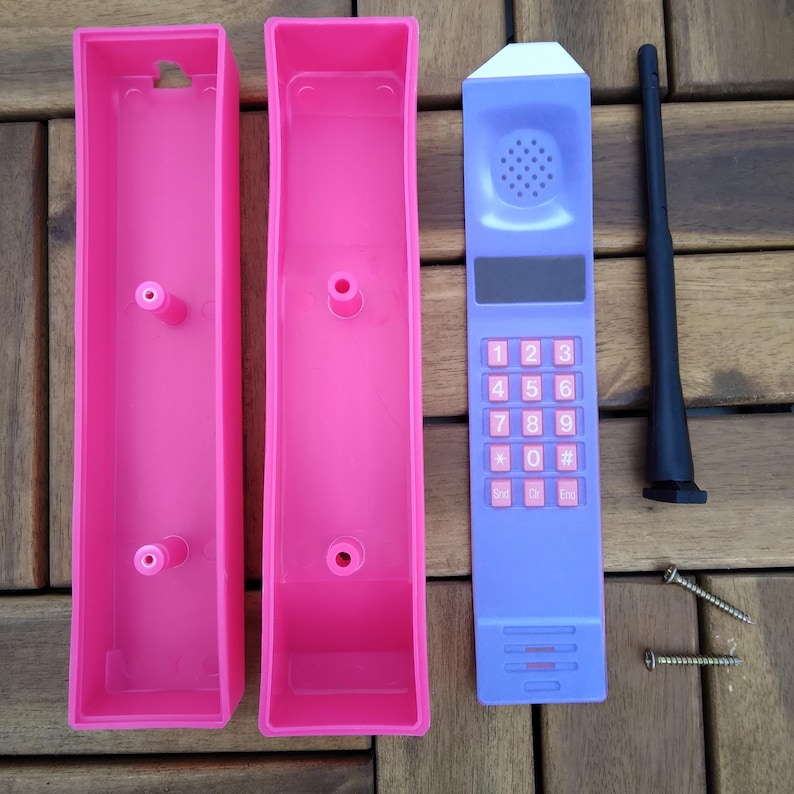 May include: A disassembled toy cellular phone with a purple keypad and antenna, pink casing, and two screws. The phone has a white top, a speaker, and a number pad with pink buttons. The pink casing is in two parts.