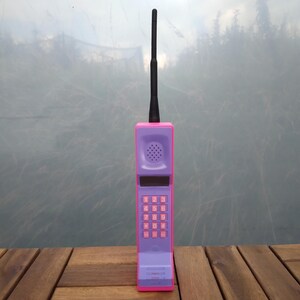 May include: A retro-style toy cell phone in purple and pink, with a black antenna. The phone features a keypad with pink buttons and a speaker. The design is reminiscent of early mobile phones from the 1980s and 1990s.