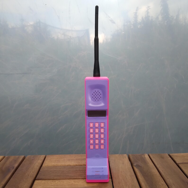 May include: A retro-style toy cell phone in purple and pink, with a black antenna. The phone features a keypad with pink buttons and a speaker. The design is reminiscent of early mobile phones from the 1980s and 1990s.