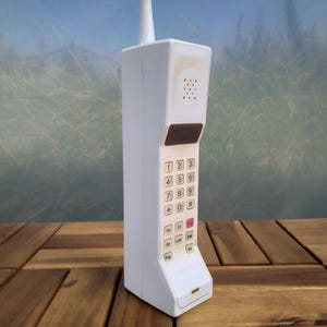 May include: A white vintage cell phone with a black keypad and a retractable antenna. The phone has a small screen and several buttons, including "Red", "Send", "End", "Stop", "Lock", "Edit", "Pager", and "Vol".