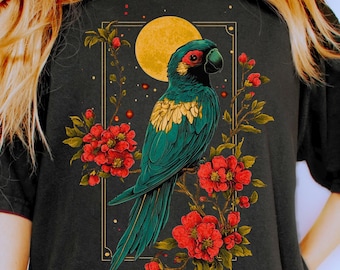 May include: A black short-sleeve t-shirt featuring a graphic print of a teal parrot perched on a branch with red flowers and a yellow moon. The design is framed by a thin gold rectangular border, creating a detailed illustration on the front.