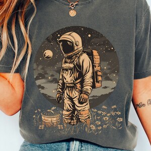 May include: A dark grey t-shirt featuring a graphic of an astronaut standing in a field of flowers. The astronaut is wearing a white spacesuit and a helmet. The background is a dark blue sky with stars and a moon.
