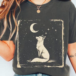May include: Graphite-coloured t-shirt with a cream illustration of a fox looking at a crescent moon and stars, framed in a rectangle. The shirt is worn with blue jeans and white trainers.