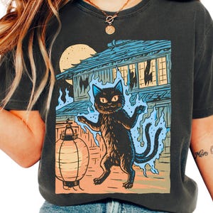 May include: A charcoal gray short-sleeve t-shirt featuring a graphic print of a two-tailed black cat surrounded by blue flames, standing next to a round paper lantern in front of a traditional building under a large yellow moon.