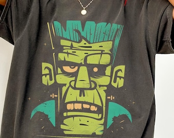 Puede incluir: Una camiseta de manga corta gris carbón con un gran estampado gráfico estilizado de una cara de monstruo verde con peinado de corte plano, ceja poblada y mandíbula prominente, utilizando tonos de verde, negro y crema en el diseño.