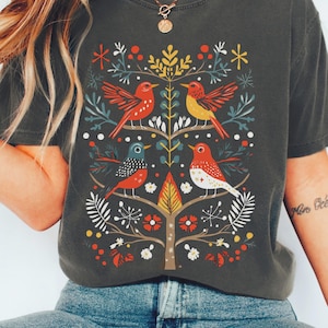 Peut inclure: Un t-shirt gris anthracite à manches courtes avec un imprimé graphique central représentant quatre oiseaux stylisés perchés sur un arbre avec des branches, des feuilles et des baies dans des tons de rouge, jaune, bleu et blanc. Col rond.