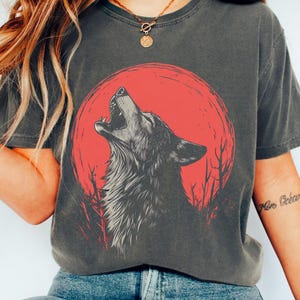 May include: A charcoal grey t-shirt featuring a graphic print of a wolf howling at a red moon. The wolf is silhouetted against the moon and the background is a dark forest.