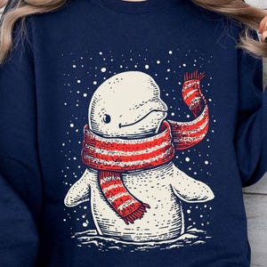 May include: A navy blue crewneck sweatshirt featuring a graphic illustration of a white beluga whale wearing a red and white striped scarf. The whale is depicted against a dark background with white dots representing falling snow.