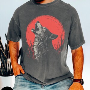 May include: A dark gray t-shirt with a graphic of a wolf howling at a red moon. The wolf is in the center of the design, with the moon behind it. The moon is a bright red color, and the wolf is a dark gray color. The t-shirt is a relaxed fit and has a crew neck.