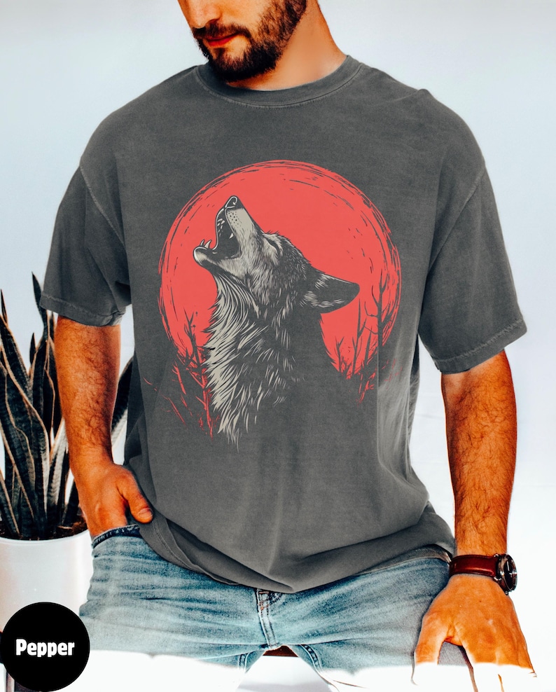May include: A dark gray t-shirt with a graphic of a wolf howling at a red moon. The wolf is in the center of the design, with the moon behind it. The moon is a bright red color, and the wolf is a dark gray color. The t-shirt is a relaxed fit and has a crew neck.