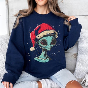Könnte beinhalten: Marineblaues Sweatshirt mit einem Cartoon-Alien, das einen roten Weihnachtsmannhut trägt. Das Alien hat große rote Augen und ist von Schneeflocken umgeben. Das Wort "Navy" ist auf einem schwarzen Kreis aufgedruckt.