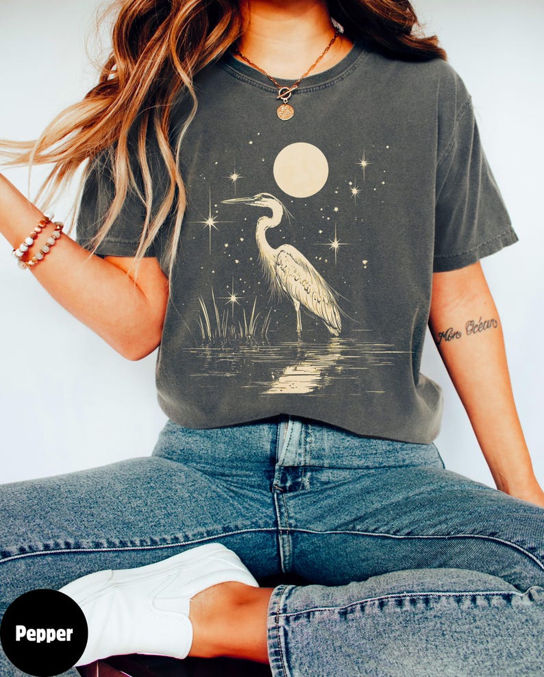 Puede incluir: Una camiseta gris oscuro con una garza blanca de pie en el agua bajo una luna llena y estrellas. La camiseta tiene un aspecto desgastado y est&aacute; hecha de una tela suave y c&oacute;moda.