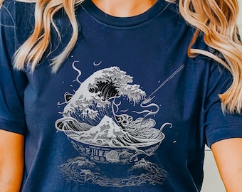 May include: A navy blue t-shirt featuring a graphic of a large wave crashing into a bowl of ramen noodles. The wave is styled to resemble the Great Wave off Kanagawa, a famous Japanese woodblock print. The text on the bowl reads "Japanese Green Tea" and "Ramen Shop".