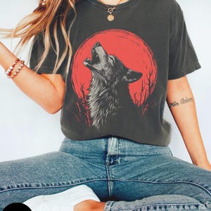 May include: A dark gray t-shirt with a graphic of a wolf howling at a red moon. The wolf is black and white and the moon is red. The t-shirt has a relaxed fit and short sleeves.