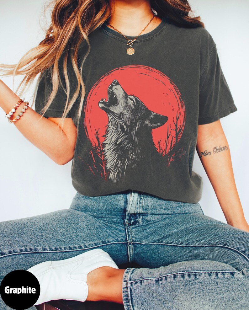 May include: A dark gray t-shirt with a graphic of a wolf howling at a red moon. The wolf is black and white and the moon is red. The t-shirt has a relaxed fit and short sleeves.