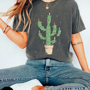 以下が含まれることがあります： 暗いグレーのTシャツに、クリスマスの電飾をつけた緑色のサボテンが描かれています。サボテンは茶色の鉢植えで、赤と黄色の電飾が付いています。Tシャツは使い古したような風合いになっており、袖には「メリークリスマス」の文字が書かれています。