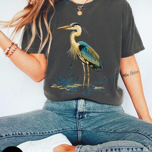 May include: A graphite grey t-shirt featuring a graphic of a blue heron standing in a marsh. The heron has a long neck and long legs. The t-shirt is made from a soft, comfortable material.