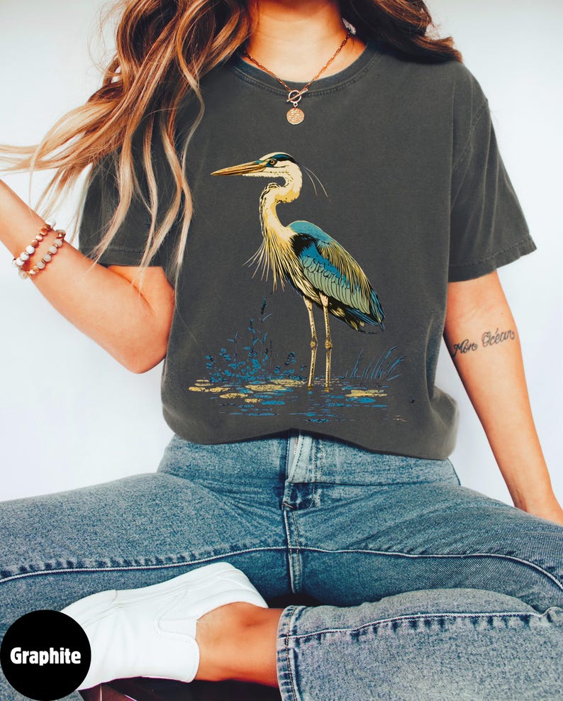 May include: A graphite grey t-shirt featuring a graphic of a blue heron standing in a marsh. The heron has a long neck and long legs. The t-shirt is made from a soft, comfortable material.