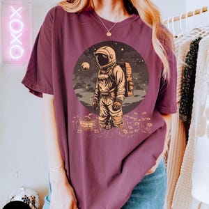 May include: A maroon t-shirt with a graphic of an astronaut standing in a field of flowers. The astronaut is wearing a white spacesuit and a backpack. The background is a starry night sky with a planet in the distance.