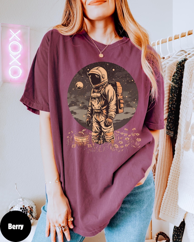 May include: A maroon t-shirt with a graphic of an astronaut standing in a field of flowers. The astronaut is wearing a white spacesuit and a backpack. The background is a starry night sky with a planet in the distance.
