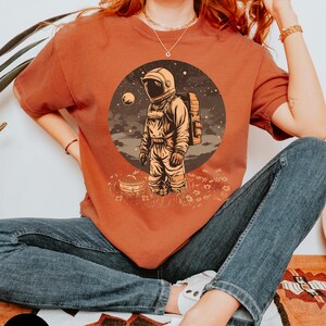 May include: A woman wearing a rust-colored t-shirt with a graphic of an astronaut standing on a planet with a moon in the background. The astronaut is wearing a white helmet and a space suit. The graphic is surrounded by a circle of stars.