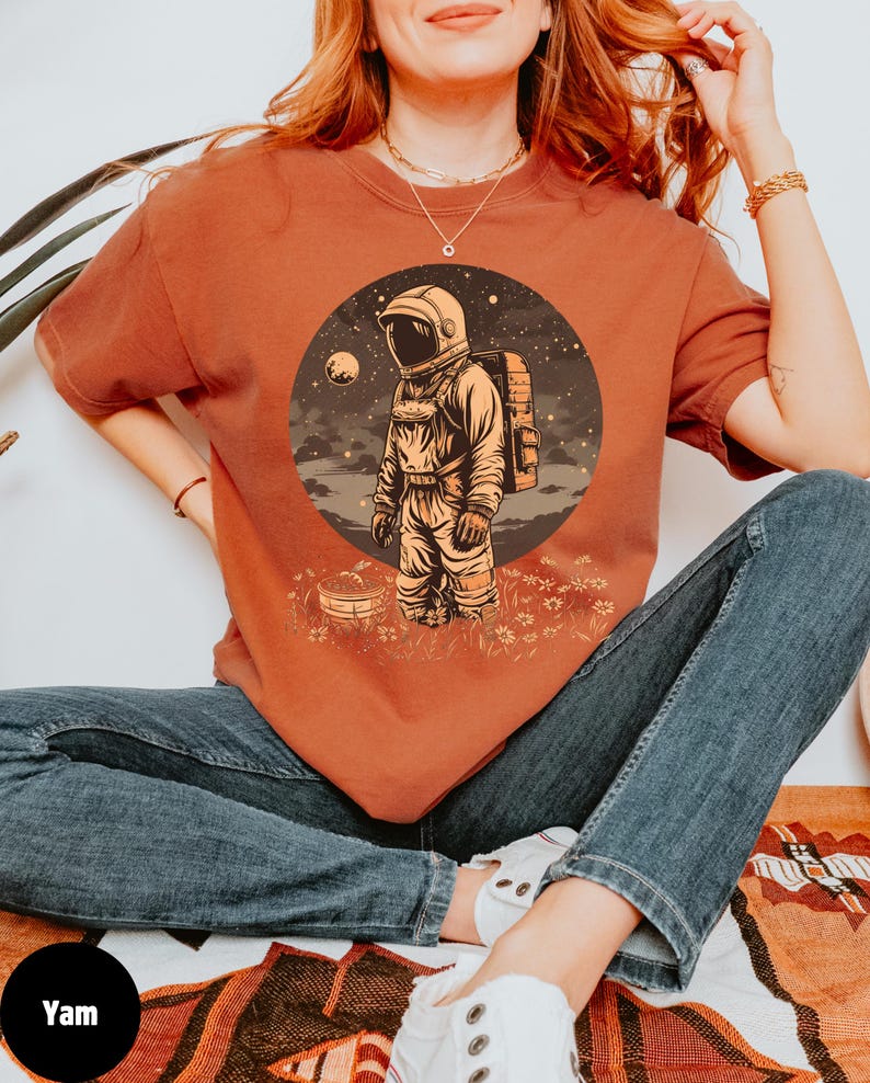 May include: A woman wearing a rust-colored t-shirt with a graphic of an astronaut standing on a planet with a moon in the background. The astronaut is wearing a white helmet and a space suit. The graphic is surrounded by a circle of stars.