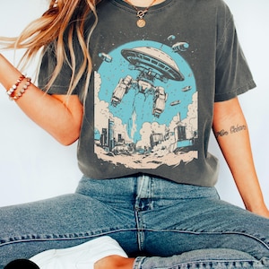 May include: A dark grey t-shirt featuring a graphic print of a spaceship hovering over a city skyline. The spaceship is blue and white with a large, round, saucer-shaped top. The city skyline is in the background and is made up of tall buildings. The graphic is in a retro style.