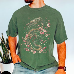 Pu&ograve; includere: Una maglietta verde con una stampa grafica di un drago dorato con fiori di ciliegio rosa. Il drago &egrave; avvolto attorno a un ramo con fiori rosa e petali che cadono dal ramo.
