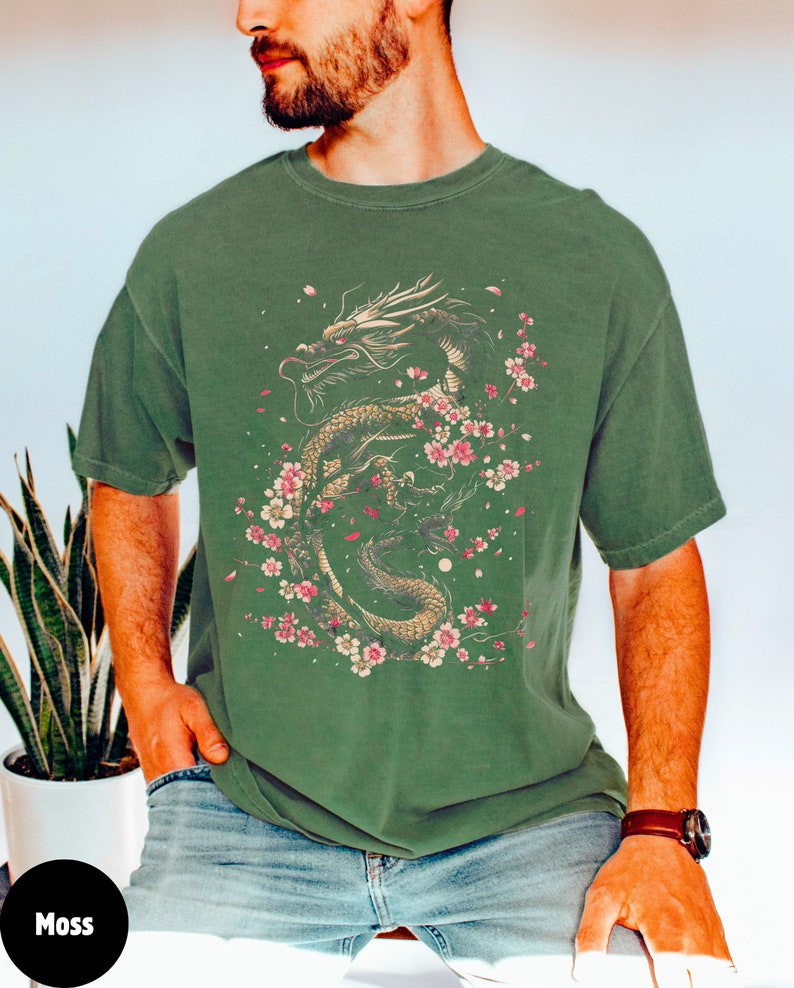 Pu&ograve; includere: Una maglietta verde con una stampa grafica di un drago dorato con fiori di ciliegio rosa. Il drago &egrave; avvolto attorno a un ramo con fiori rosa e petali che cadono dal ramo.
