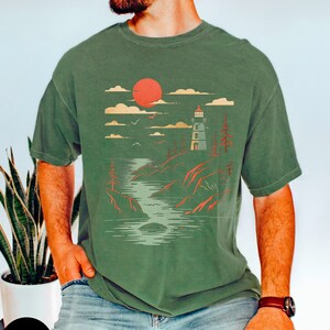 May include: A green t-shirt with a graphic print of a lighthouse, a red sun, and a body of water. The graphic is in a vintage style.