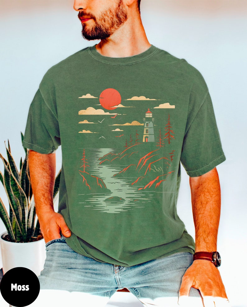 May include: A green t-shirt with a graphic print of a lighthouse, a red sun, and a body of water. The graphic is in a vintage style.