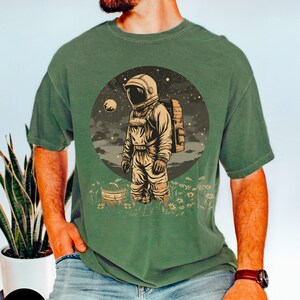 May include: A green t-shirt with a vintage illustration of an astronaut standing in a field of flowers. The astronaut is wearing a white helmet and a spacesuit. The background is a dark blue sky with stars and a moon.