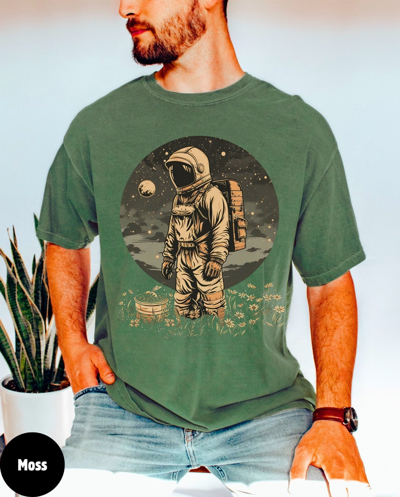 May include: A green t-shirt with a vintage illustration of an astronaut standing in a field of flowers. The astronaut is wearing a white helmet and a spacesuit. The background is a dark blue sky with stars and a moon.