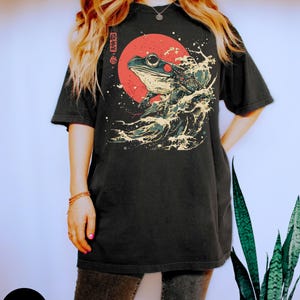 Pode incluir: Camiseta preta com um gr&aacute;fico de um sapo numa onda com um sol vermelho no fundo. Os caracteres japoneses na camiseta dizem "onda de sapo".