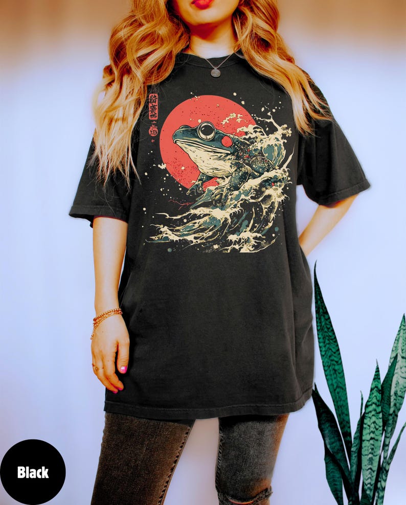 Pode incluir: Camiseta preta com um gr&aacute;fico de um sapo numa onda com um sol vermelho no fundo. Os caracteres japoneses na camiseta dizem "onda de sapo".