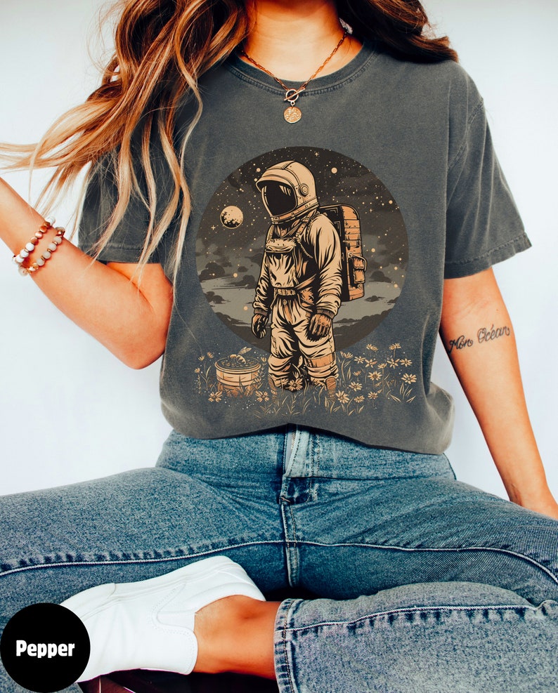 May include: A dark grey t-shirt featuring a graphic of an astronaut standing in a field of flowers. The astronaut is wearing a white spacesuit and a helmet. The background is a dark blue sky with stars and a moon.
