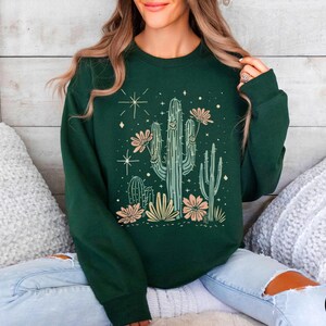 May include: A forest green sweatshirt with a graphic print of a cactus with flowers and stars. The sweatshirt has long sleeves and a crew neck.