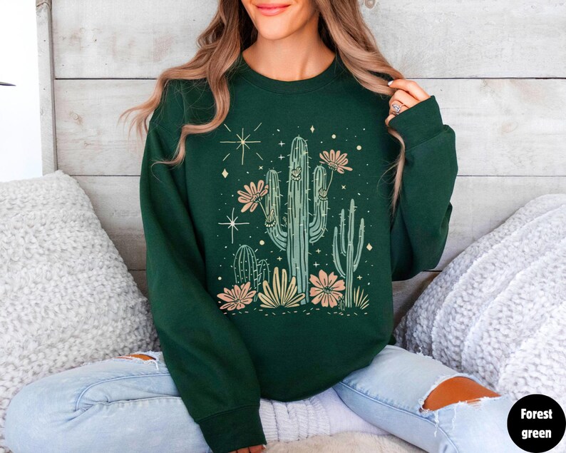 May include: A forest green sweatshirt with a graphic print of a cactus with flowers and stars. The sweatshirt has long sleeves and a crew neck.