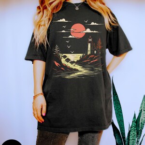 May include: A black t-shirt with a graphic of a lighthouse on a rocky shore with a red sun setting behind it. The graphic is in a vintage style with a yellow and orange color scheme.