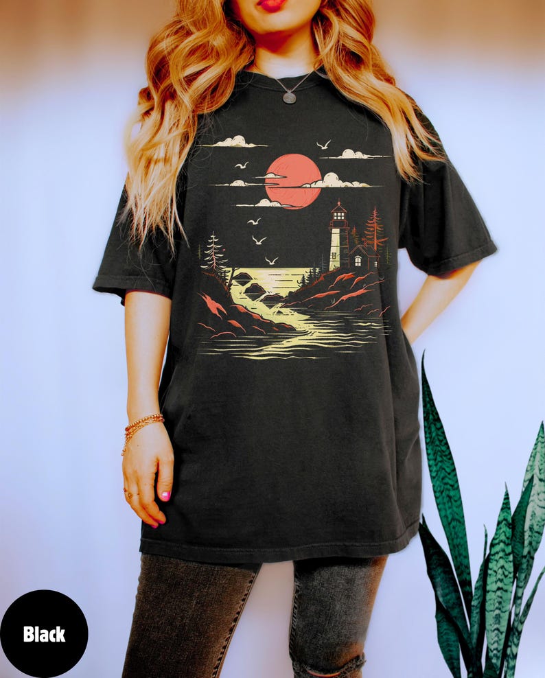 May include: A black t-shirt with a graphic of a lighthouse on a rocky shore with a red sun setting behind it. The graphic is in a vintage style with a yellow and orange color scheme.
